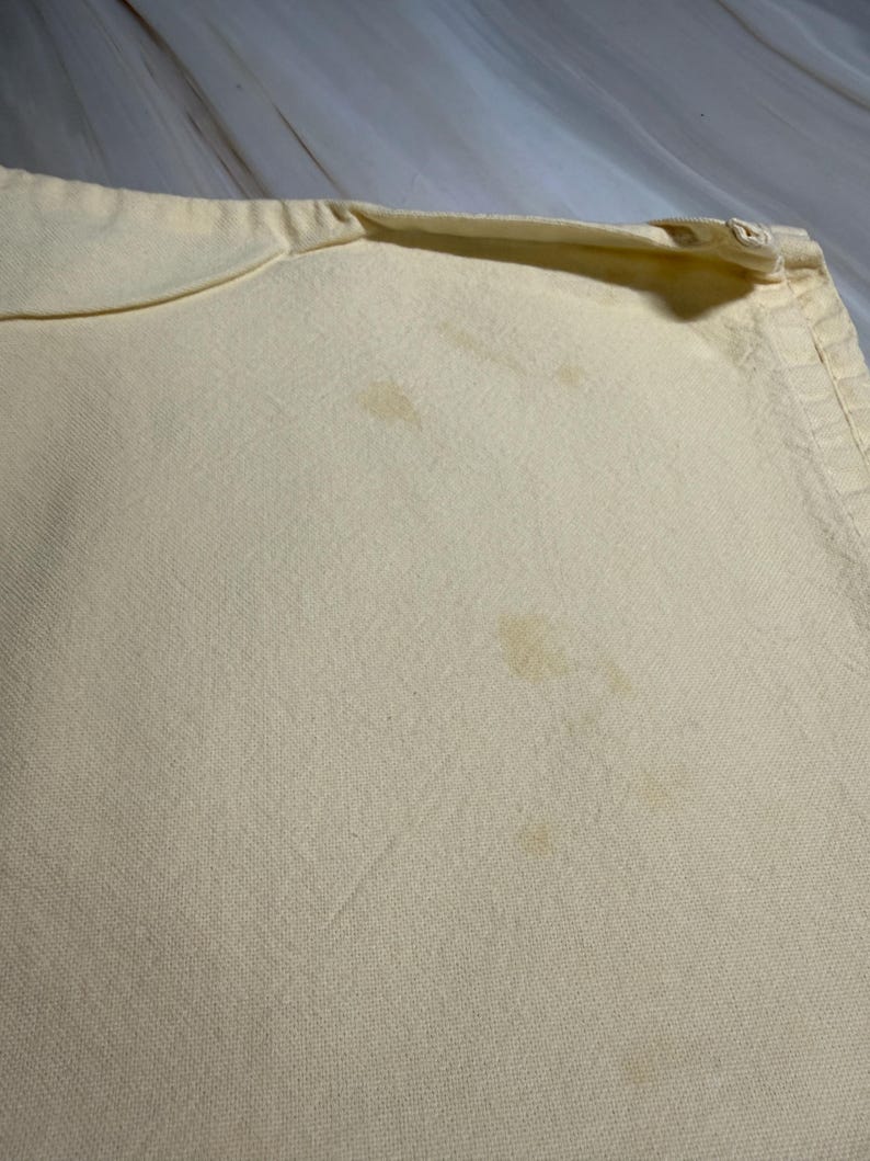 Op de afbeelding: Close-up van een lichtgele kussensloop met zichtbare vlekken. De kussensloop heeft een ritssluiting aan &eacute;&eacute;n kant en een licht gebogen rand. De stof lijkt een geweven materiaal te zijn.