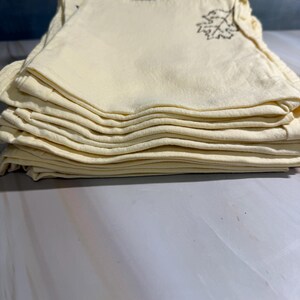 Op de afbeelding: Een stapel gevouwen, lichtgele stoffen servetten. Elke servet heeft een bruin bladontwerp geborduurd in de hoek. De servetten zijn netjes opgestapeld en tonen de gevouwen randen en de geborduurde details.