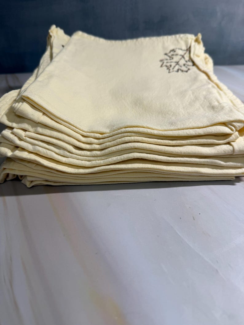 Op de afbeelding: Een stapel gevouwen, lichtgele stoffen servetten. Elke servet heeft een bruin bladontwerp geborduurd in de hoek. De servetten zijn netjes opgestapeld en tonen de gevouwen randen en de geborduurde details.
