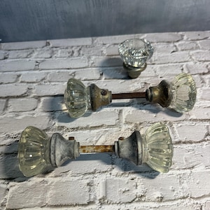 May include: Three vintage glass door knobs with metal hardware. The knobs have a ribbed design and are clear. The metal components show signs of age and wear, with a rusty appearance. The knobs are arranged on a white brick background.