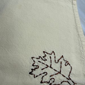 Op de afbeelding: Cr&egrave;mekleurige linnen servet met een bruin geborduurd bladontwerp. Het blad is een gedetailleerde omtrek, die een vleugje herfst aan het textiel toevoegt. De textuur van de servet is zichtbaar, wat wijst op een zacht, absorberend materiaal.