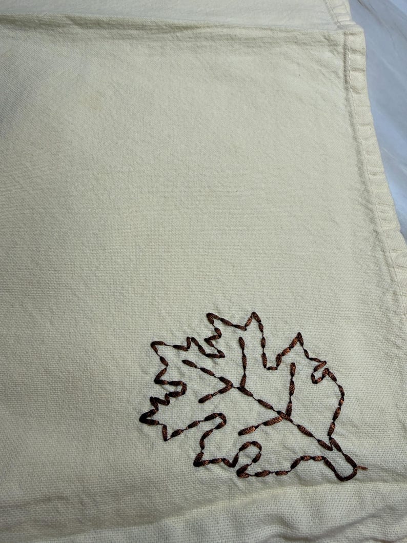 Op de afbeelding: Cr&egrave;mekleurige linnen servet met een bruin geborduurd bladontwerp. Het blad is een gedetailleerde omtrek, die een vleugje herfst aan het textiel toevoegt. De textuur van de servet is zichtbaar, wat wijst op een zacht, absorberend materiaal.