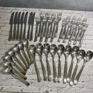 May include: A collection of stainless steel silverware, including knives, forks, and spoons, arranged on a white brick surface. The silverware has a shiny, reflective finish, and the arrangement suggests a formal dining setting.