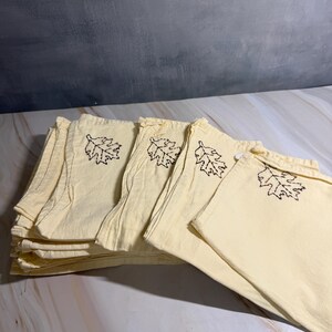 Op de afbeelding: Een stapel lichtgele stoffen servetten met een donkerbruin geborduurd bladmotief. De servetten zijn gevouwen en opgestapeld, met meerdere zichtbaar. De achtergrond is een gevlekte grijs-witte ondergrond.