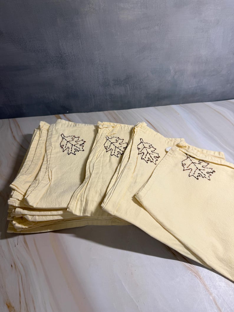 Op de afbeelding: Een stapel lichtgele stoffen servetten met een donkerbruin geborduurd bladmotief. De servetten zijn gevouwen en opgestapeld, met meerdere zichtbaar. De achtergrond is een gevlekte grijs-witte ondergrond.