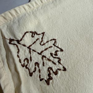 Op de afbeelding: Cr&egrave;mekleurige stof met een gedetailleerd bruin geborduurd bladmotief. Het blad is gecentreerd op de stof en toont de textuur van het borduurwerk. De stof heeft een subtiele, natuurlijke tint.