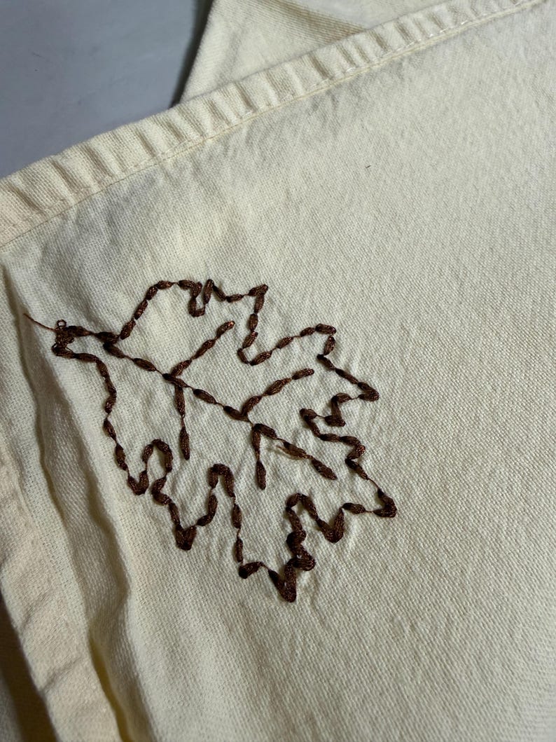 Op de afbeelding: Cr&egrave;mekleurige stof met een gedetailleerd bruin geborduurd bladmotief. Het blad is gecentreerd op de stof en toont de textuur van het borduurwerk. De stof heeft een subtiele, natuurlijke tint.