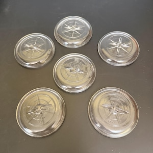 May include: Six clear glass compass-themed coasters. Each coaster features a compass rose design in the center, with directional lines radiating outwards. The coasters are round and have a slightly raised edge.