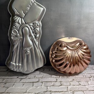 May include: Two vintage metal cake molds. One is a silver-colored, detailed mold in the shape of a woman. The other is a copper-colored, round mold shaped like a seashell. Both molds have a hole for hanging.