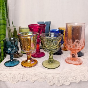 May include: A collection of multi-colored glass goblets featuring various textures and patterns. The set includes individual pieces in shades of deep blue, amber, magenta, olive green, and soft pink, each with a distinct stem and base design.