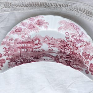 May include: A crescent-shaped ceramic dish featuring a white base with a detailed pink transferware pattern. The design depicts a landscape with a house, a bridge, a sailboat, and floral elements. The dish has a scalloped edge along the outer rim.