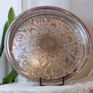 May include: A large, round, silver-toned metal serving tray featuring an intricate, embossed floral and scrollwork pattern across its surface. The tray has a raised, decorative rope-style border and a smooth, polished center.
