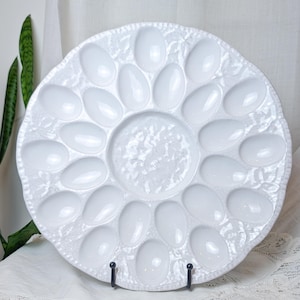 May include: A round white ceramic egg serving platter featuring a central circular compartment surrounded by eighteen oval-shaped indentations. The surface of the platter has a textured, embossed leaf pattern and a scalloped edge.