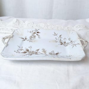 May include: A rectangular white ceramic serving tray featuring brown floral and leaf illustrations across the surface. The tray includes two integrated handles on the sides with matching brown decorative patterns and molded corner details.