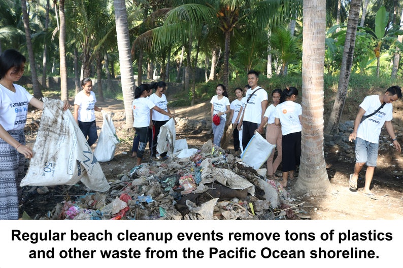 May include: A group of people wearing white t-shirts with text are cleaning up trash on a beach. They are holding large bags and removing waste from a pile of debris. The scene is set on a sunny day with palm trees in the background.