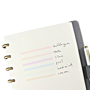 May include: A disc-bound notebook with a dark gray cover and gold-toned discs, featuring a white page with six horizontal highlighter swatches in pink, peach, lilac, blue, mint, and tan, labeled with handwritten text including bubblegum and cocoa.