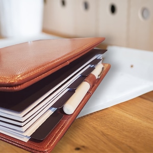 May include: A brown leather bound journal with multiple dividers and a white pen. The journal is open and the pages are visible.
