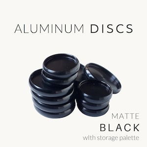 May include: A stack of matte black aluminum discs with a storage palette. The text reads ALUMINUM DISCS, MATTE BLACK, and with storage palette. The discs are circular with a raised rim and a smooth, dark finish, arranged in two small piles.