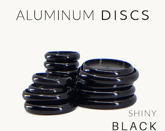 May include: A collection of shiny black aluminum discs stacked in two piles, accompanied by a storage palette. The text reads ALUMINUM DISCS, SHINY BLACK, and with storage palette. The discs have a smooth, reflective finish and circular shape.