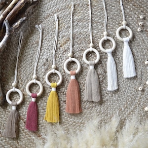 Può includere: Sette ciondoli per auto in legno a forma di anello con nappe di vari colori, tra cui beige, bianco, giallo, arancione, rosso e marrone. I ciondoli sono appesi a cordini bianchi e sono esposti su una superficie intrecciata.