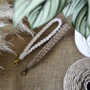 Peut inclure: Deux lanières de poignet en macramé, une crème et une beige avec des accents dorés, sont posées sur une surface en toile de jute. Chaque lanière a un fermoir doré. L'arrière-plan comprend de l'herbe de la pampa séchée et du feuillage vert.