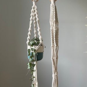Pode incluir: Dois suportes de plantas de macramé com anéis de madeira. O suporte à esquerda tem uma planta verde em um vaso branco e azul. Ambos os suportes são feitos de cordão de algodão branco.