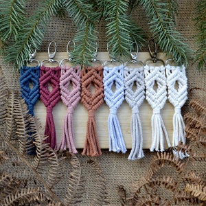 Puede incluir: Siete llaveros de macramé de colores con borlas que cuelgan de una superficie de madera. Los llaveros son en tonos de azul, rojo, rosa, marrón, azul claro y blanco.