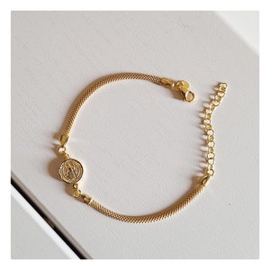 May include: Gold-toned bracelet featuring a round pendant with a detailed religious emblem. The bracelet has a mesh-like chain design and an adjustable chain for a comfortable fit. The clasp and pendant are also gold-toned.