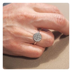 May include: A silver ring with a round, detailed charm featuring a cross and the words "Ave Maria" and "Gratia Plena".