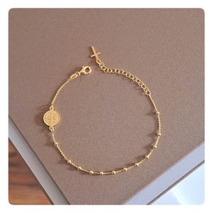 May include: A gold-colored bracelet featuring a small cross charm and a circular pendant with a cross design. The bracelet has a delicate chain with small, evenly spaced beads. The clasp is a lobster claw style.