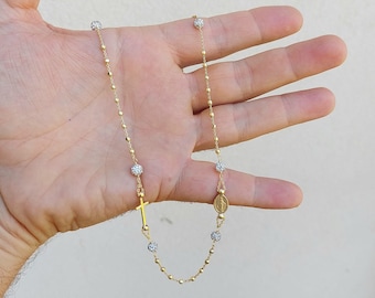 May include: A gold chain necklace with a small cross pendant and a round charm with a religious image. The chain is adorned with small, round beads that have a clear crystal-like appearance.