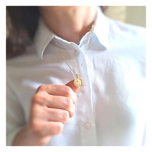 Può includere: Una collana con catena d'oro con un piccolo ciondolo rotondo. Il ciondolo ha un design in rilievo.
