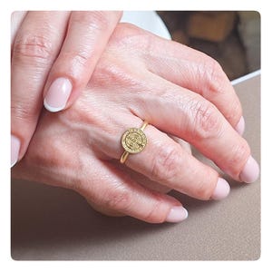 May include: A gold ring with a round, detailed design. The ring is worn on a finger with a light pink nail polish.
