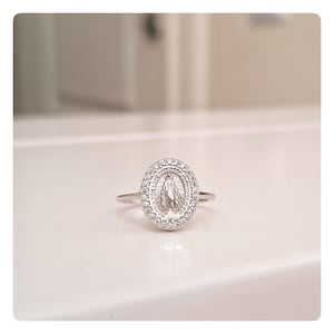 Peut inclure: Une bague en argent avec un pendentif ovale représentant une figure religieuse, entourée de petites pierres claires. La bague est posée sur une surface blanche, mettant en valeur son design et ses détails.