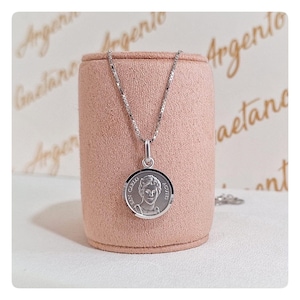 Puede incluir: Un collar de plata con un colgante redondo que presenta un retrato y el texto "SAN CARLO ACUTIS". El colgante cuelga de una delicada cadena. El collar se exhibe sobre un soporte cilíndrico rosa.