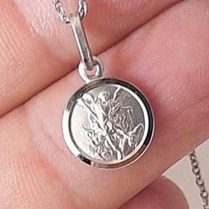 May include: A silver pendant necklace featuring a round charm with a detailed relief of a saint slaying a dragon. The pendant is attached to a delicate chain. The image is a close-up, highlighting the intricate design.