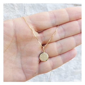 May include: A gold-colored necklace featuring a small, round pendant with an intricate design. The pendant hangs from a delicate chain. The necklace is displayed against a light background.