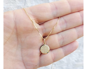 Könnte beinhalten: Eine goldfarbene Halskette mit einem kleinen, runden Anhänger mit einem aufwendigen Design. Der Anhänger hängt an einer zarten Kette. Die Halskette wird vor einem hellen Hintergrund gezeigt.