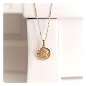 Pode incluir: Um colar com pingente dourado com um medalhão circular com um retrato e texto. O medalhão é suspenso por uma delicada corrente de ouro. O pingente tem o texto "PADRE PIO DA PIETRELCINA" e o ano "1910".