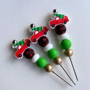 Peut inclure: Trois décorations de sapin de Noël en forme de camion rouge avec des perles vertes et blanches et des perles dorées. Les camions transportent des sapins de Noël et ont un motif à carreaux rouge et noir.