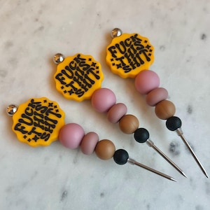 May include: Three cocktail picks with yellow toppers that read "FUCK THIS SHIT" in black. The picks have pink, brown, and black beads, and a silver tip.