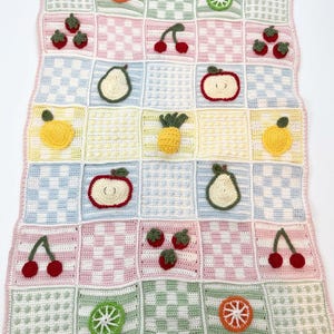 May include: A rectangular crochet blanket featuring a grid of squares in pastel colours. The squares alternate between chequered patterns, textured dots, and various crocheted fruit appliques including cherries, strawberries, pears, apples, pineapples, and citrus slices.