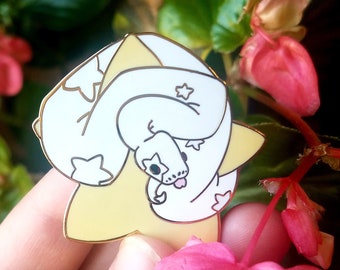 May include: A white snake pin with star-shaped markings and a pink tongue, coiled on a yellow star-shaped base. The pin has a gold border. The background includes pink flowers and green foliage.
