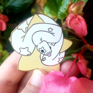 Peut inclure: Épingle à nourrice en forme de serpent blanc avec des étoiles et une langue rose, posée sur une base en forme d'étoile jaune. Le fond est composé de fleurs roses et de feuilles vertes.