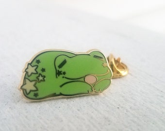 May include: A green enamel pin featuring a sleeping frog with star accents. The frog has a light pink belly and a gold-colored outline. The pin is a cute accessory.
