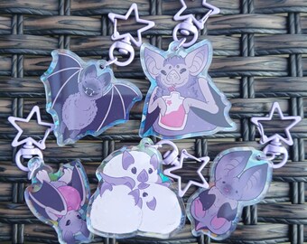 May include: Five acrylic keychains featuring illustrated bats in various poses, including one drinking from a pink pouch, one hanging upside down, and one white fluffy bat. Each charm has a light purple star-shaped metal swivel clasp.