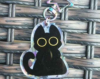 May include: A black cat acrylic keychain featuring large yellow circular eyes and a holographic background effect. The charm is attached to a star-shaped metal clasp with an iridescent rainbow finish, providing a reflective and colorful appearance.