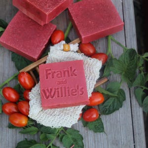 Può includere: Diverse saponette quadrate rosse, una delle quali presenta il testo in rilievo Frank and Willie's, sono disposte con pomodorini rossi freschi e tralci di pomodoro verdi su una superficie di legno. Un piccolo sacchetto a rete intrecciato color crema si trova sotto il sapone.
