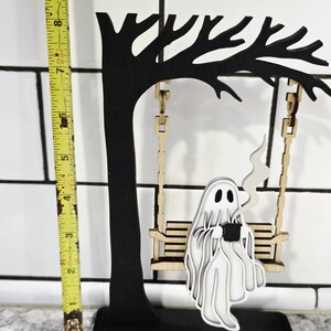 May include: A black wooden tree with a swing, holding a layered white ghost figure. The ghost is holding a black cup with steam rising. The swing is made of light wood. The tree is approximately 8 inches tall.