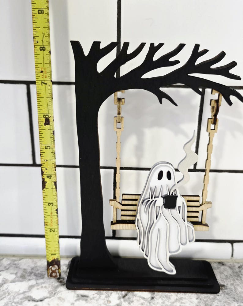 May include: A black wooden tree with a swing, holding a layered white ghost figure. The ghost is holding a black cup with steam rising. The swing is made of light wood. The tree is approximately 8 inches tall.